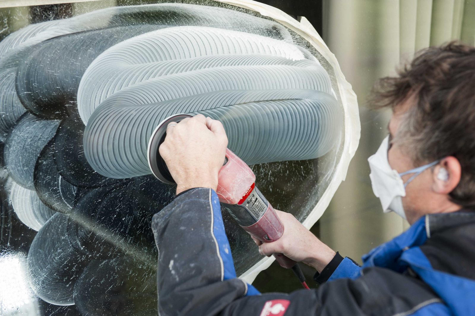 Technieker met stofmasker en veiligheidsbril houdt een polijstkop tegen een grote glaspartij; het glas is nat en vertoont cirkelvormige polijstsporen