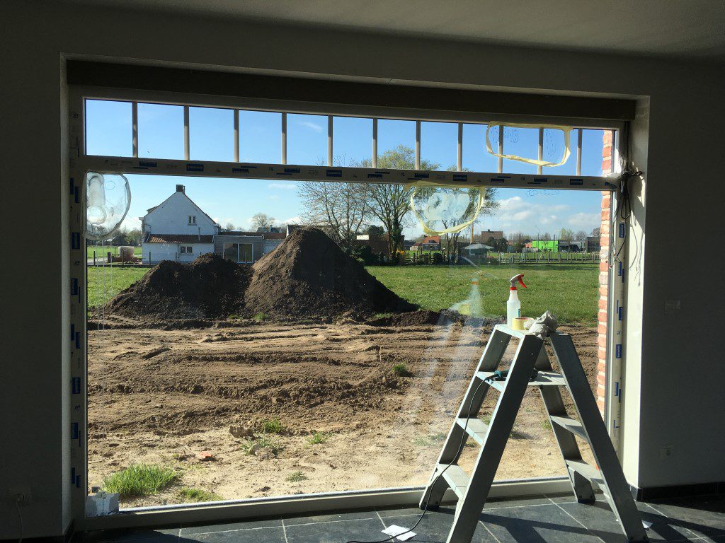 Bouwwerf gezien door een raam: grondhoop, ladder en bouwmateriaal vlak achter het glas