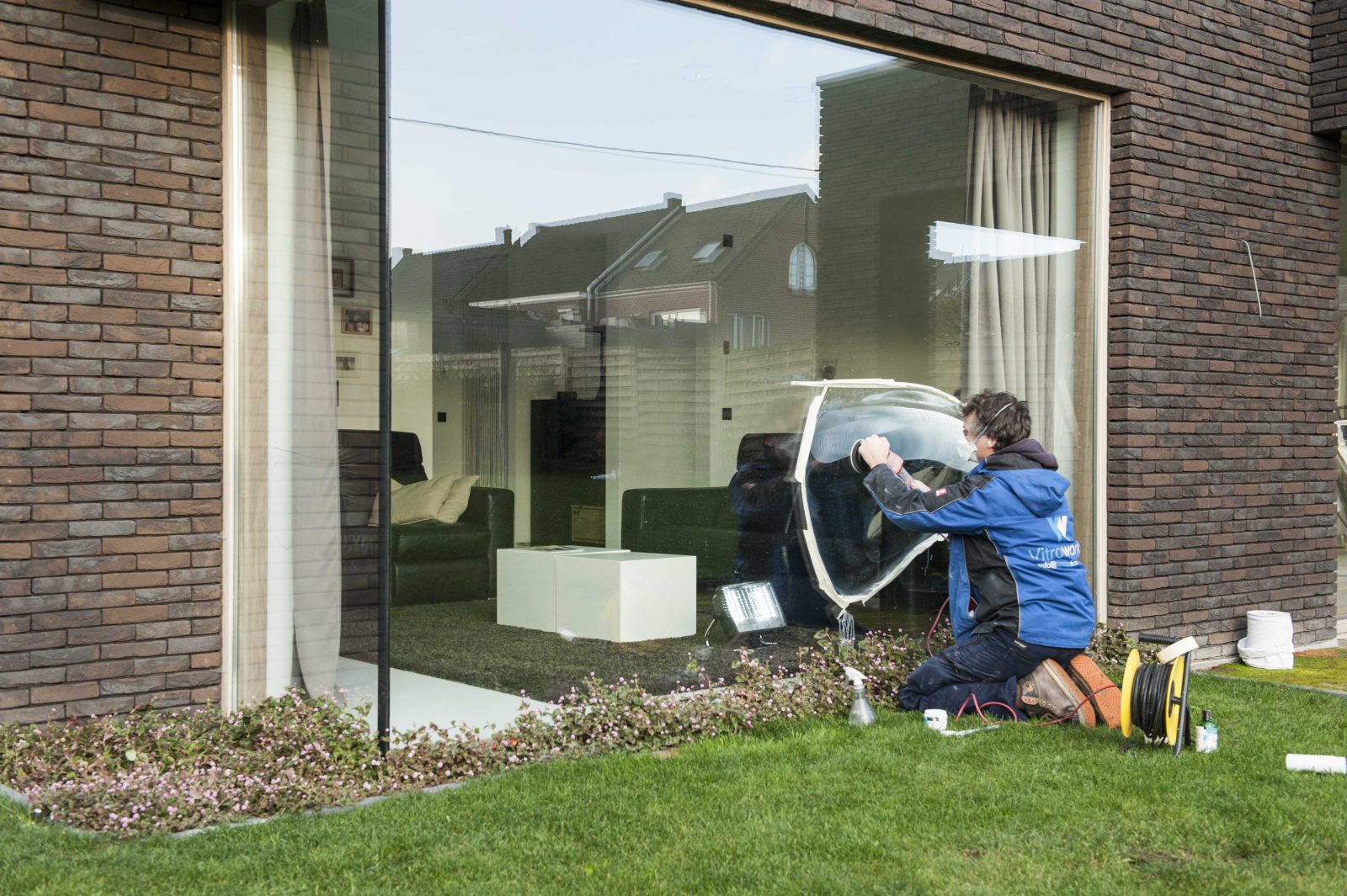 Technieker geknield aan de grote raampartij van een woning, met haspel en materiaal op het gazon naast zich